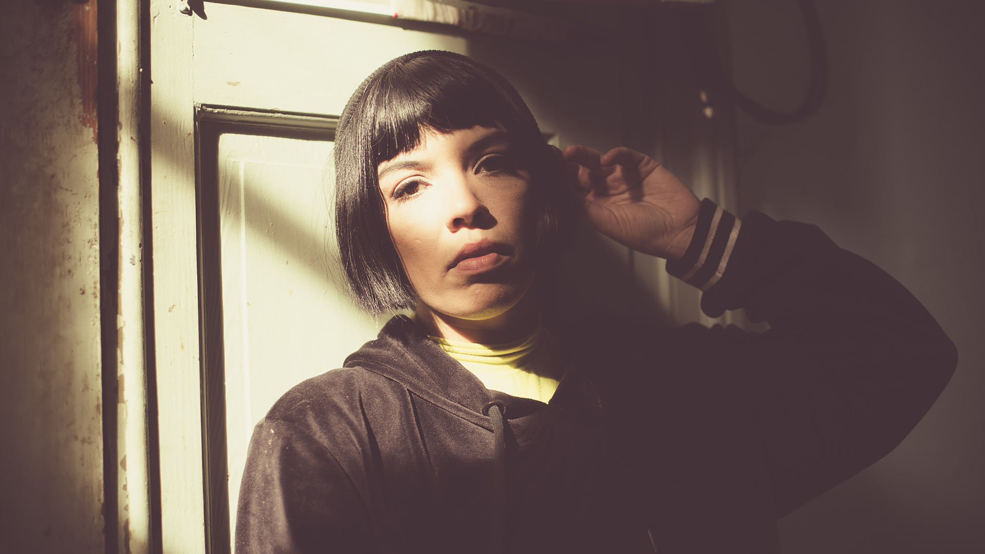 A person with a dark bob haircut, a key look for autumn hair trends, wears a black hoodie over a yellow top whilst positioned against a light-coloured door.