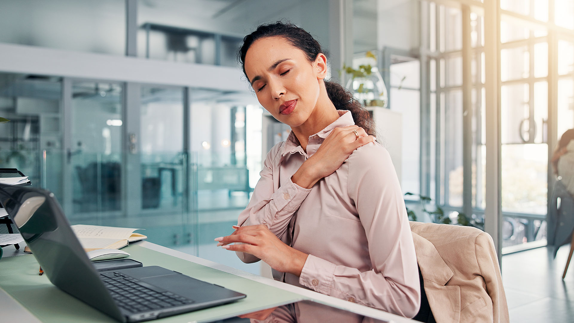 A woman sitting at a desk with a laptop, holding her shoulder and grimacing in pain caused by poor posture.